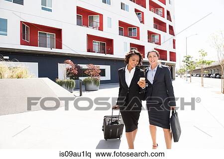 Stock Photography - Two businesswomen walking outdoors, pulling suitcase. Fotosearch