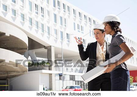 Stock Photo - Two businesswomen, wearing hard hats, carrying plans. Fotosearch