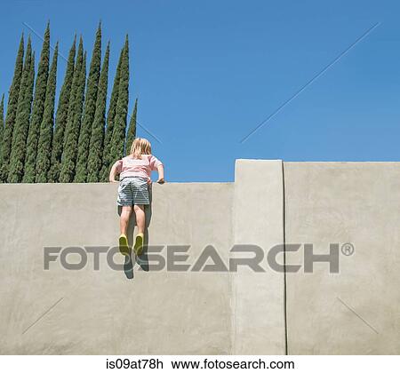 Stock Image - Young boy looking over tall wall, feet dangling, rear view. Fotosearch
