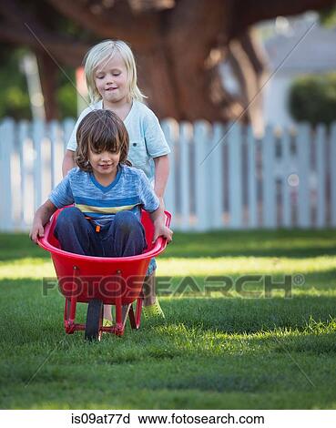 Stock Image - Young boy sitting in wheelbarrow, his friend pushing him. Fotosearch