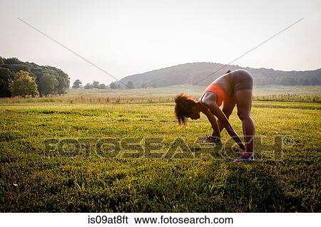 Stock Photograph - Young female runner touching toes in rural park. Fotosearch