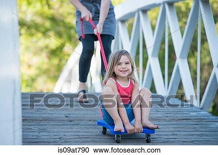 Stock Image - Young girl sitting on wheeled board, mother helping her roll over wooden bridge. Fotosearch