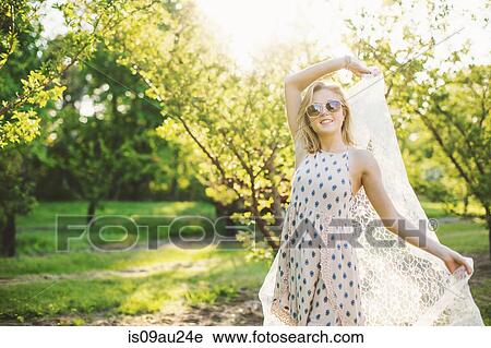 Young woman in orchard wearing sleeveless dress and sunglasses holding lace fabric, looking at camera smiling View Large Photo Image Stock Image - Young woman in orchard wearing sleeveless dress and sunglasses holding lace fabric, looking at camera smiling. Fotosearch