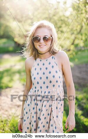 Stock Image - Young woman wearing sleeveless dress and sunglasses looking at camera smiling. Fotosearch