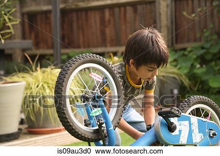 Stock Image - Boy in garden repairing upside down bicycle looking down. Fotosearch