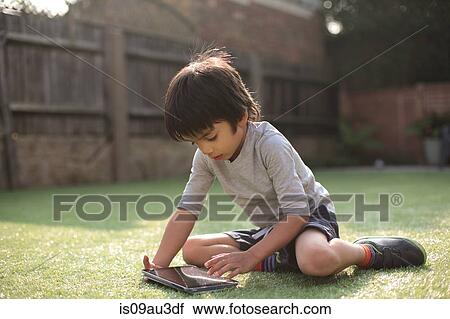 Stock Image - Boy in garden sitting on grass looking down using digital tablet. Fotosearch