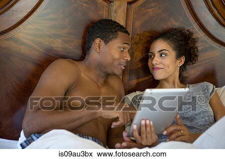 Stock Photo - Couple in bed using digital tablet face to face smiling. Fotosearch