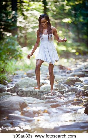 Stock Photograph - Glamorous young woman wearing white dress stepping across forest river rocks. Fotosearch