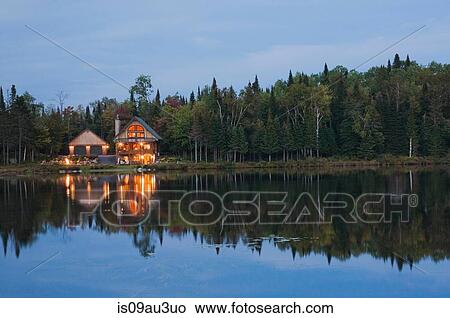 Picture - Illuminated garage and handcrafted spruce log home on edge of lake at dusk, Quebec, Canada. Fotosearch