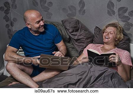 Male couple sitting up in bed laughing whilst reading and using laptop View Large Photo Image Stock Photograph - Male couple sitting up in bed laughing whilst reading and using laptop. Fotosearch