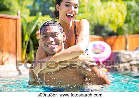 Stock Photography - Mid adult man carrying young woman on back in swimming pool looking at camera smiling. Fotosearch