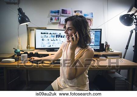 Stock Image - Mid adult woman at desk, using smartphone. Fotosearch
