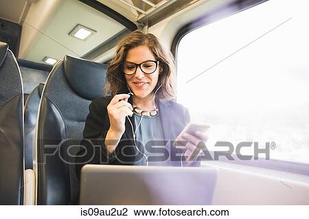 Stock Image - Mid adult woman on train, holding smartphone, looking at laptop. Fotosearch