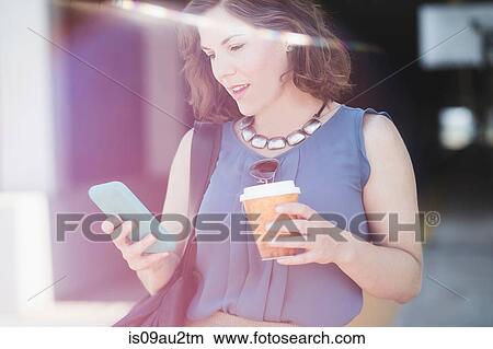 Mid adult woman outdoors, using smartphone, holding coffee cup View Large Photo Image Picture - Mid adult woman outdoors, using smartphone, holding coffee cup. Fotosearch