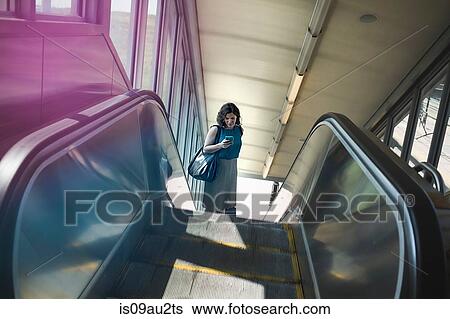 Stock Photograph - Mid adult woman using escalator, holding smartphone, elevated view. Fotosearch