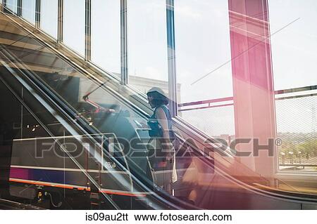 Stock Photograph - Mid adult woman using escalator, holding wheeled suitcase and smartphone. Fotosearch