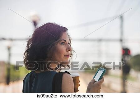 Picture - Mid adult woman waiting at train station, using smartphone, holding coffee cup. Fotosearch