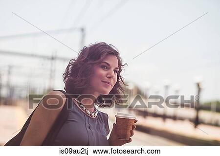 Stock Image - Mid adult woman waiting at train station, holding coffee cup. Fotosearch