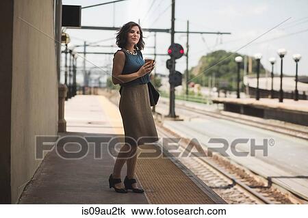 Stock Image - Mid adult woman waiting at train station, holding coffee cup. Fotosearch