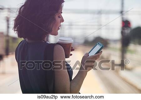 Mid adult woman waiting at train station, using smartphone, holding coffee cup View Large Photo Image Stock Photography - Mid adult woman waiting at train station, using smartphone, holding coffee cup. Fotosearch