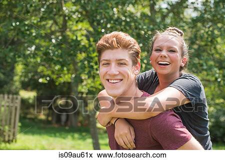 Stock Photo - Portrait of young man giving young woman piggy back looking at camera smiling. Fotosearch