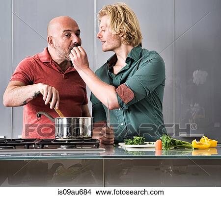Picture - Romantic male couple tasting food whilst cooking in kitchen. Fotosearch