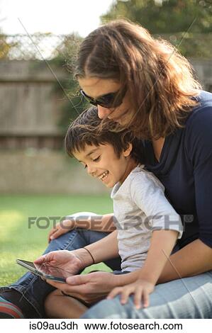 Side view of boy sitting on mother's lap using digital tablet looking down smiling View Large Photo Image Stock Image - Side view of boy sitting on mother's lap using digital tablet looking down smiling. Fotosearch