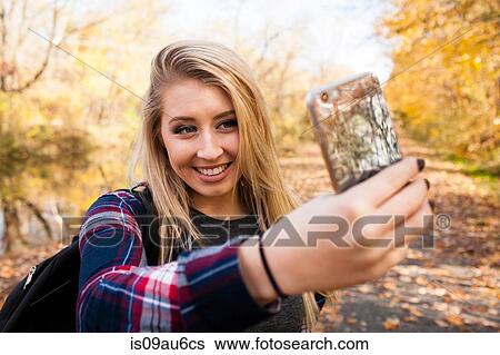 Stock Photograph - Young blond woman taking smartphone selfie in autumn forest. Fotosearch