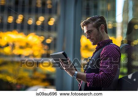 Stock Photograph - Young businessman leaning against park window reading digital tablet. Fotosearch