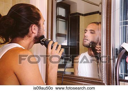 Young man looking in mirror, shaving with electric razor View Large Photo Image Picture - Young man looking in mirror, shaving with electric razor. Fotosearch