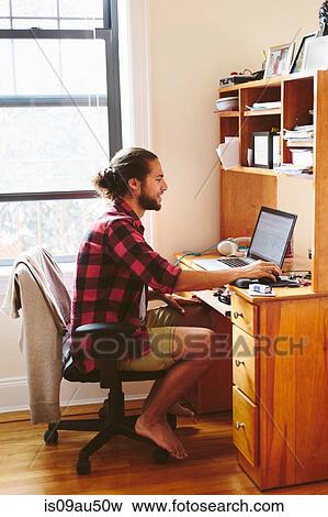 Stock Photo - Young man sitting at desk, using laptop. Fotosearch