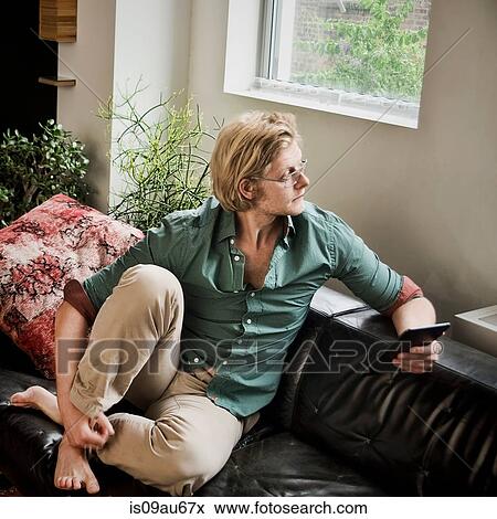 Young man sitting on sofa with digital tablet looking away View Large Photo Image Stock Photo - Young man sitting on sofa with digital tablet looking away. Fotosearch