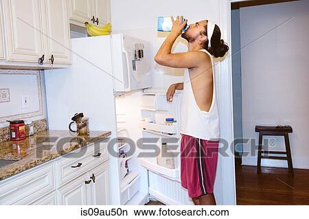 Young man standing next to open fridge, drinking milk from carton View Large Photo Image Picture - Young man standing next to open fridge, drinking milk from carton. Fotosearch