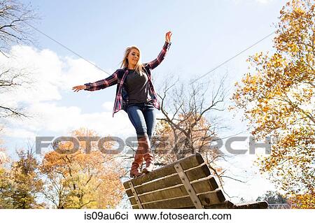 Stock Image - Young woman balancing on top of park bench in autumn forest. Fotosearch