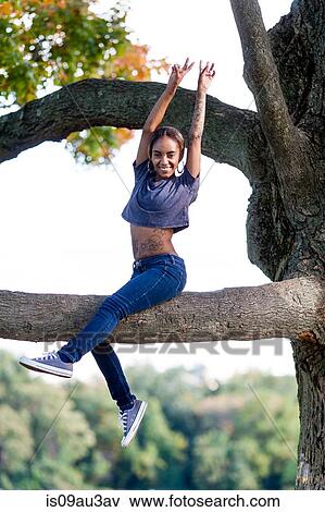 Young woman sitting in tree arms raised looking at camera smiling View Large Photo Image Stock Photo - Young woman sitting in tree arms raised looking at camera smiling. Fotosearch