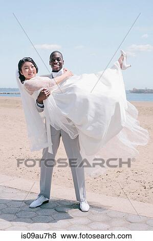 Stock Photo - Bridegroom on beach carrying bride looking at camera smiling. Fotosearch