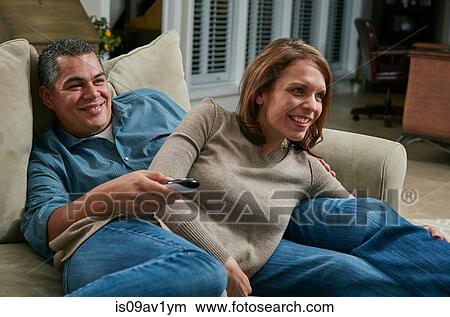 Picture - Couple snuggling on sofa, looking away using remote control smiling. Fotosearch