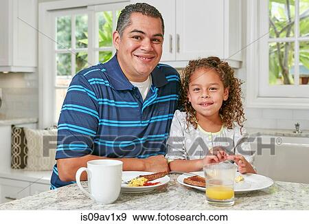 Stock Photo - Father and daughter side by side at kitchen counter eating breakfast, looking at camera smiling. Fotosearch
