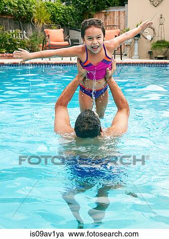 Stock Image - Father in swimming pool lifting up daughter, arms open looking at camera smiling. Fotosearch