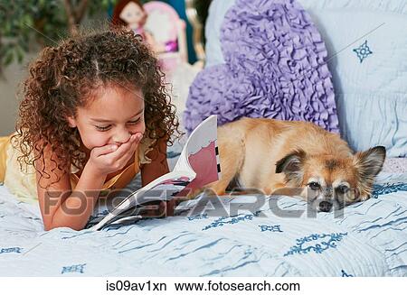 Girl and dog lying on bed reading book, hand on mouth laughing View Large Photo Image Picture - Girl and dog lying on bed reading book, hand on mouth laughing. Fotosearch