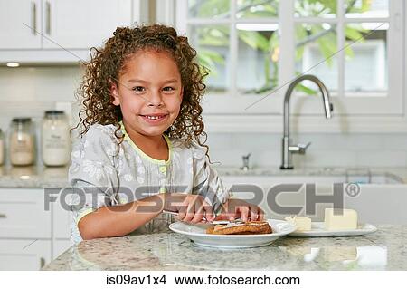 Girl at kitchen counter spreading butter on toast looking at camera smiling View Large Photo Image Picture - Girl at kitchen counter spreading butter on toast looking at camera smiling. Fotosearch