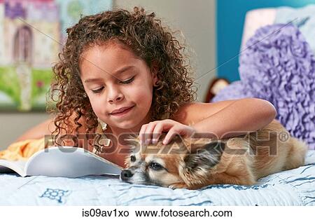 Picture - Girl lying on bed reading book stroking dog. Fotosearch