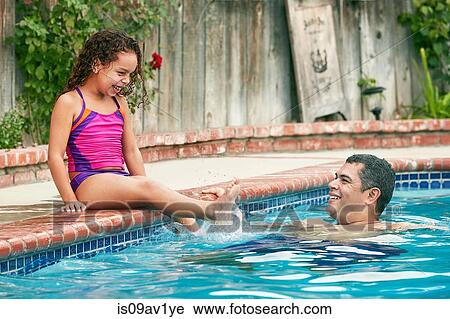 Stock Image - Girl wearing swimwear sitting poolside splashing father smiling. Fotosearch