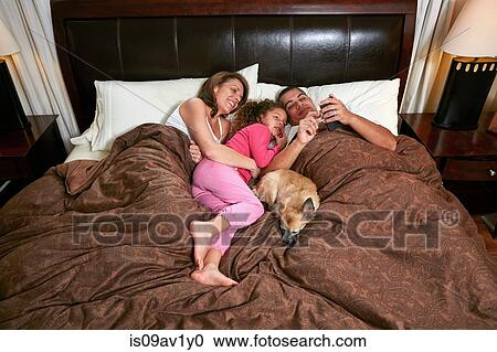 Stock Image - High angle view of girl in bed with parents and dog looking at smartphone smiling. Fotosearch