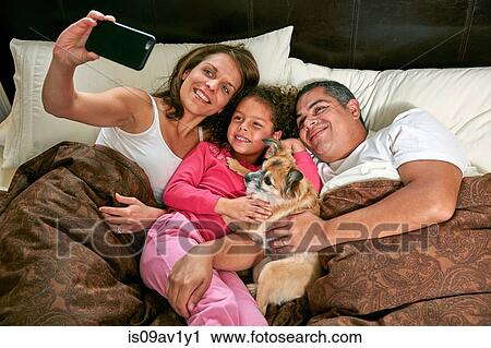 High angle view of girl in bed with parents and dog using smartphone to take selfie smiling View Large Photo Image Stock Image - High angle view of girl in bed with parents and dog using smartphone to take selfie smiling. Fotosearch