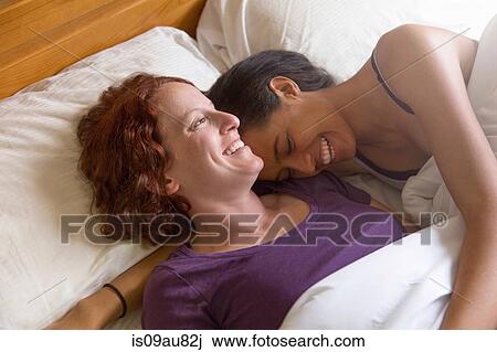 Stock Image - High angle view of lesbian couple lying in bed hugging, smiling. Fotosearch
