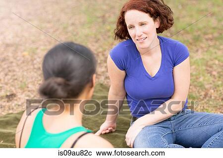 Stock Image - High angle view of young woman sitting on picnic blanket tallking to friend, smiling. Fotosearch