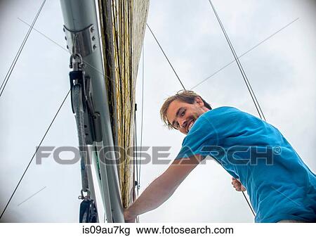 Low angle view of young man holding onto sailboat mast looking at camera smiling View Large Photo Image Stock Image - Low angle view of young man holding onto sailboat mast looking at camera smiling. Fotosearch