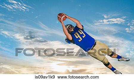 Male teenage American football player catching ball mid air against blue sky View Large Photo Image Stock Photo - Male teenage American football player catching ball mid air against blue sky. Fotosearch