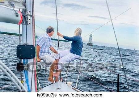 Stock Photograph - Mature couple on bow of sailboat looking away towards lighthouse. Fotosearch